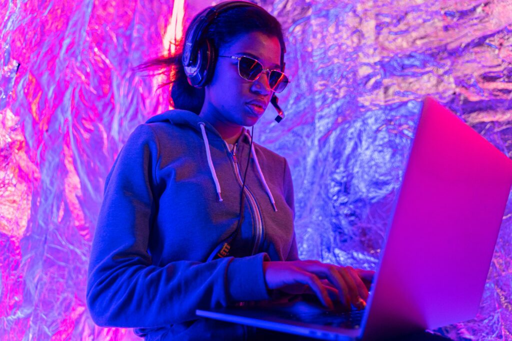 A dynamic DJ wearing sunglasses and a headset, performing with a laptop in a colorful setting.